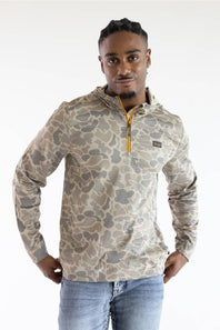 A man wearing a beige and gray camouflage quarter-zip pullover sweatshirt with a yellow zipper pull, paired with blue jeans, against a plain white studio background.