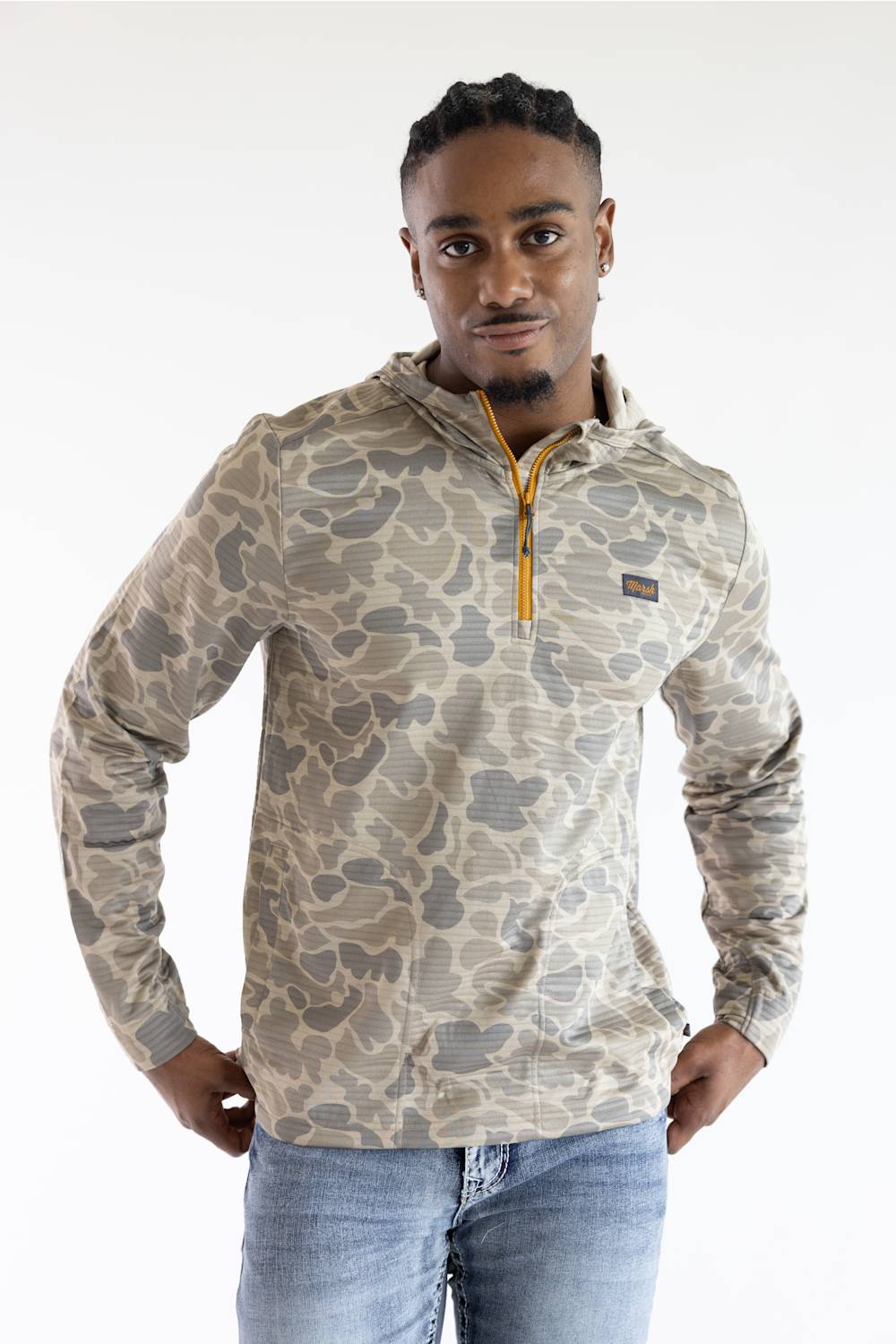 A man wearing a beige and gray camouflage quarter-zip pullover sweatshirt with a yellow zipper pull, paired with blue jeans, against a plain white studio background.