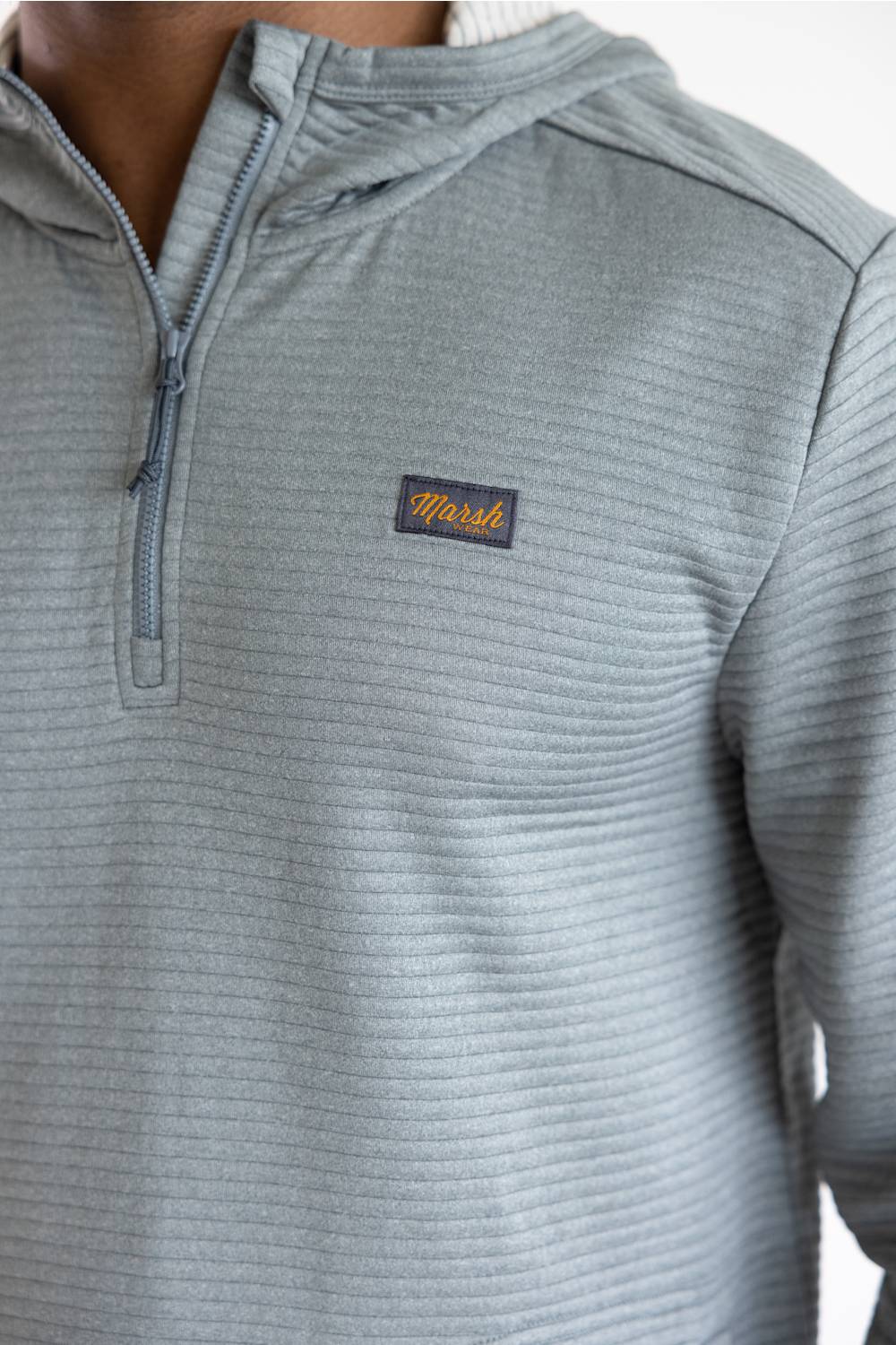 Close-up of a person wearing a light gray ribbed quarter-zip pullover with a small rectangular Marsh logo patch on the chest.