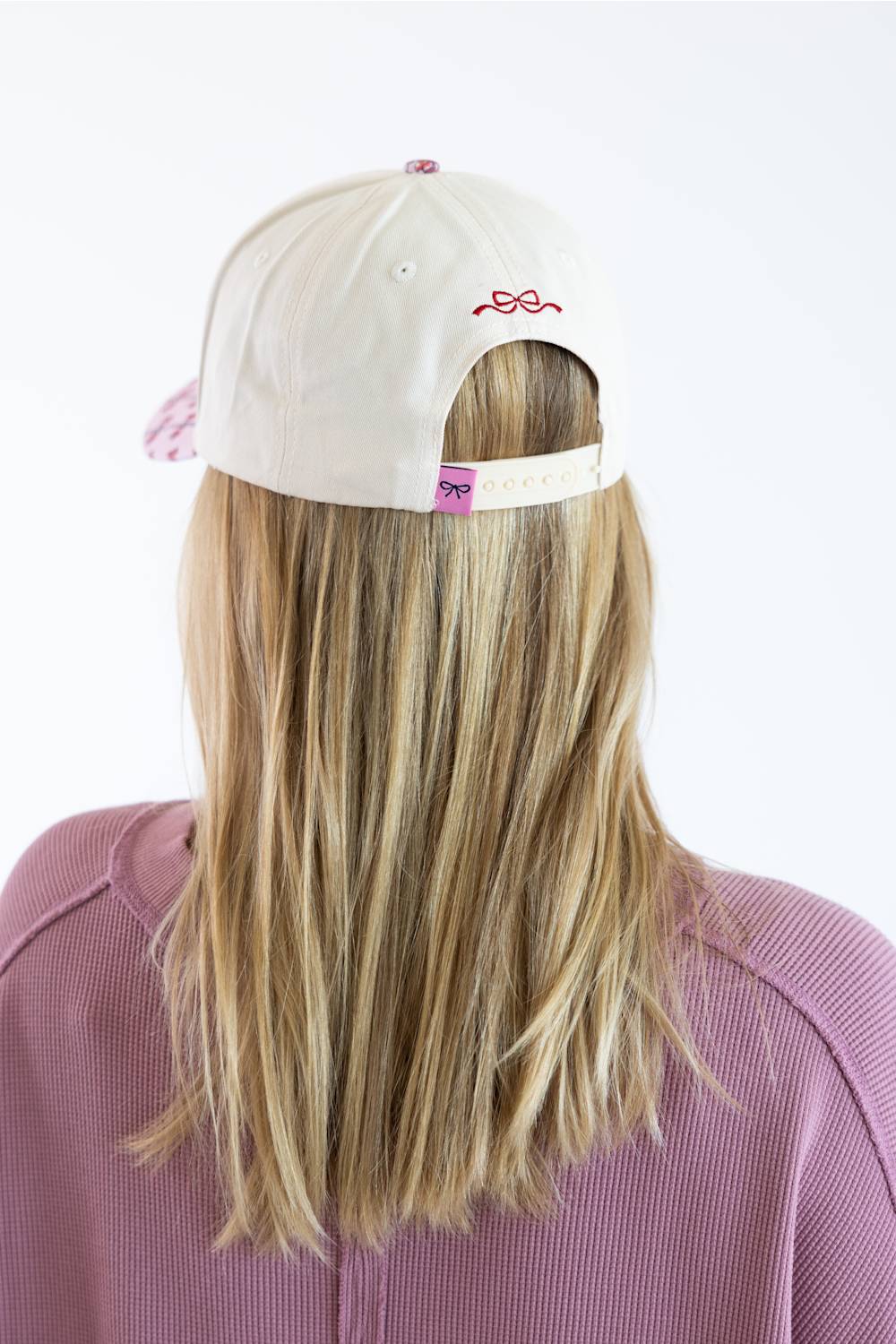 Back view of a model wearing the Simply Southern Classic Santa Holiday Hat for Women in Cream, featuring a red embroidered bow above the keyhole, a pink bow patch on the adjustable snapback closure, paired with a dusty pink sweatshirt against a plain studio background.