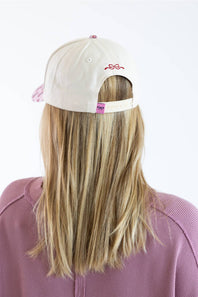 Back view of a model wearing the Simply Southern Classic Santa Holiday Hat for Women in Cream, featuring a red embroidered bow above the keyhole, a pink bow patch on the adjustable snapback closure, paired with a dusty pink sweatshirt against a plain studio background.