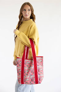 A woman wearing a yellow sweatshirt and light blue jeans holds a large tote bag with red handles and a pink and white patterned body featuring illustrated dogs.