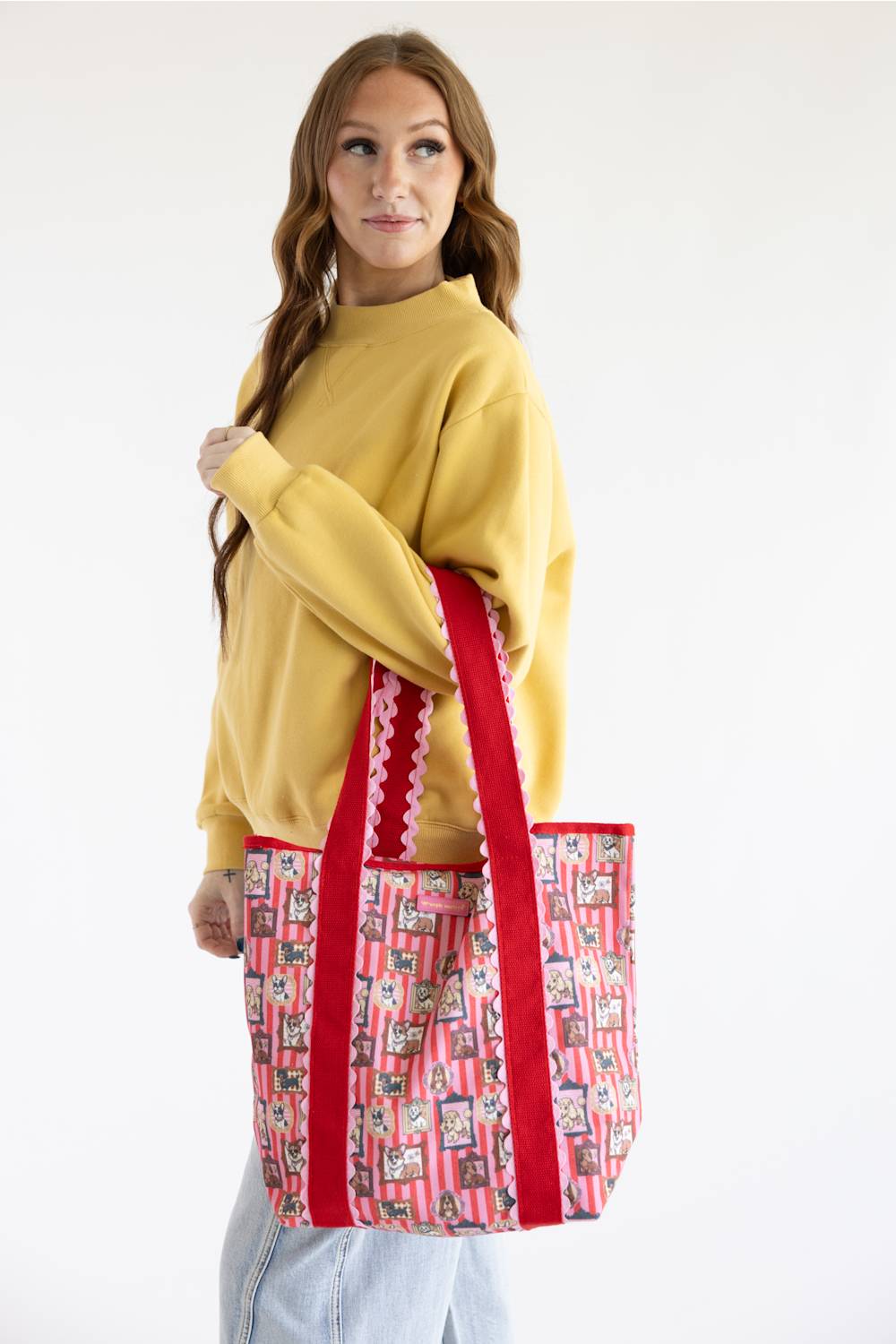A woman wearing a yellow sweatshirt and light blue jeans holds a large tote bag with red handles and a pink and white patterned body featuring illustrated dogs.