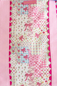 A close-up of a pink and white bag, featuring various floral, gingham, and bow patterns with pink ribbon motifs, bordered by pink satin-like trim.