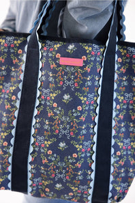 A close-up of a tote bag with a navy blue floral print, featuring scalloped light blue trim and dark navy shoulder straps. A small pink rectangular label is visible near the top center.