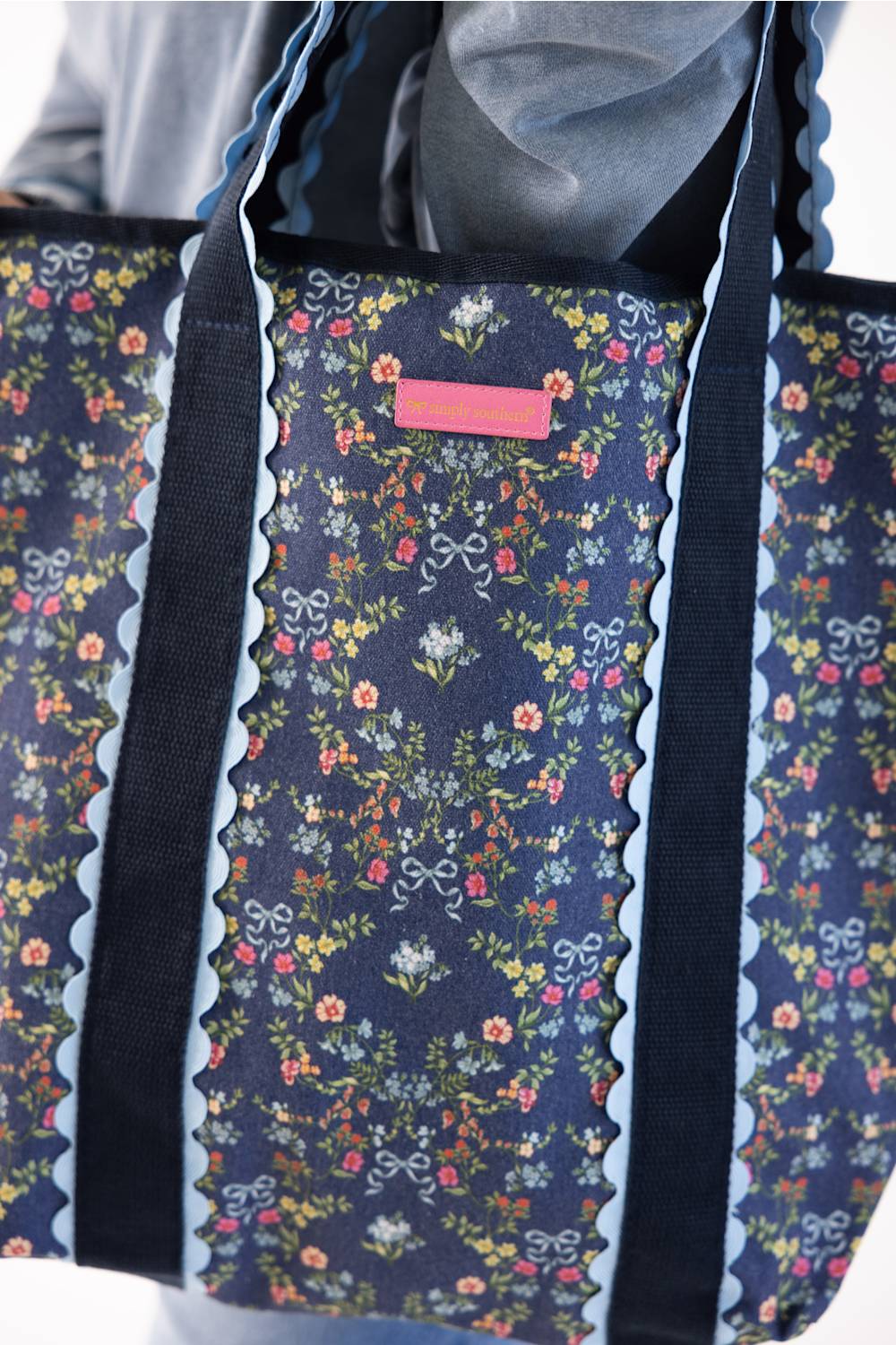 A close-up of a tote bag with a navy blue floral print, featuring scalloped light blue trim and dark navy shoulder straps. A small pink rectangular label is visible near the top center.