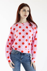 A woman modeling a pink crewneck sweatshirt with a bold red polka dot pattern, paired with blue jeans, against a plain white background.