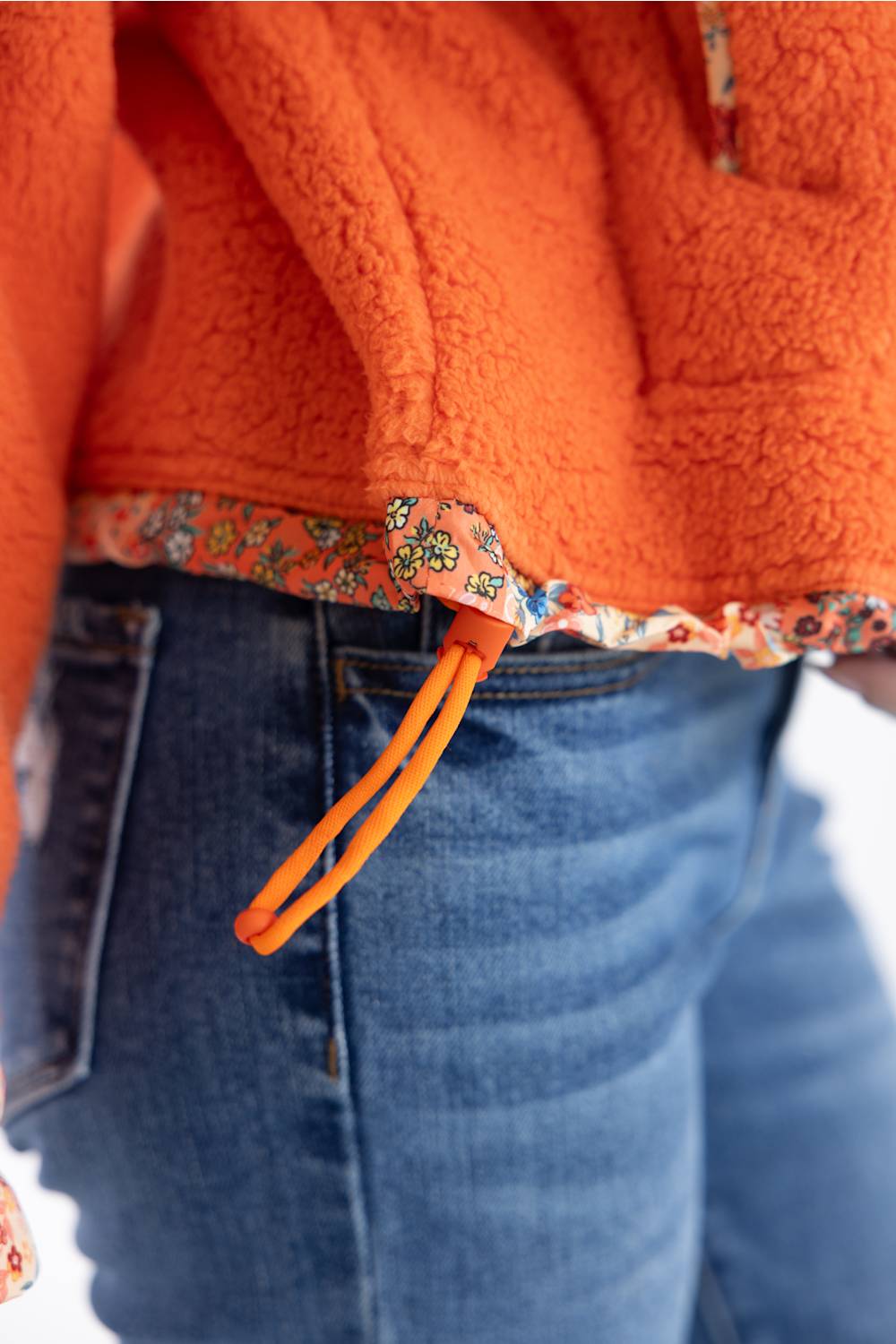 Close-up of a person wearing an orange sherpa fleece jacket with a floral-patterned hem and an orange drawstring, paired with blue jeans.