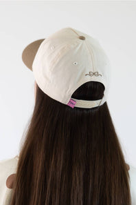A person viewed from behind wearing a cream-colored baseball cap with a brown visor and a small bow embroidery on the back, set against a plain white background.