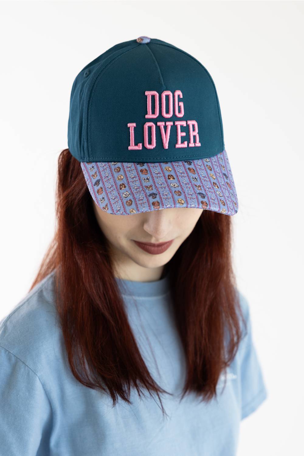 A person wearing a dark teal baseball cap with 'DOG LOVER' embroidered in pink on the front and a patterned brim, paired with a light blue t-shirt against a plain white background.