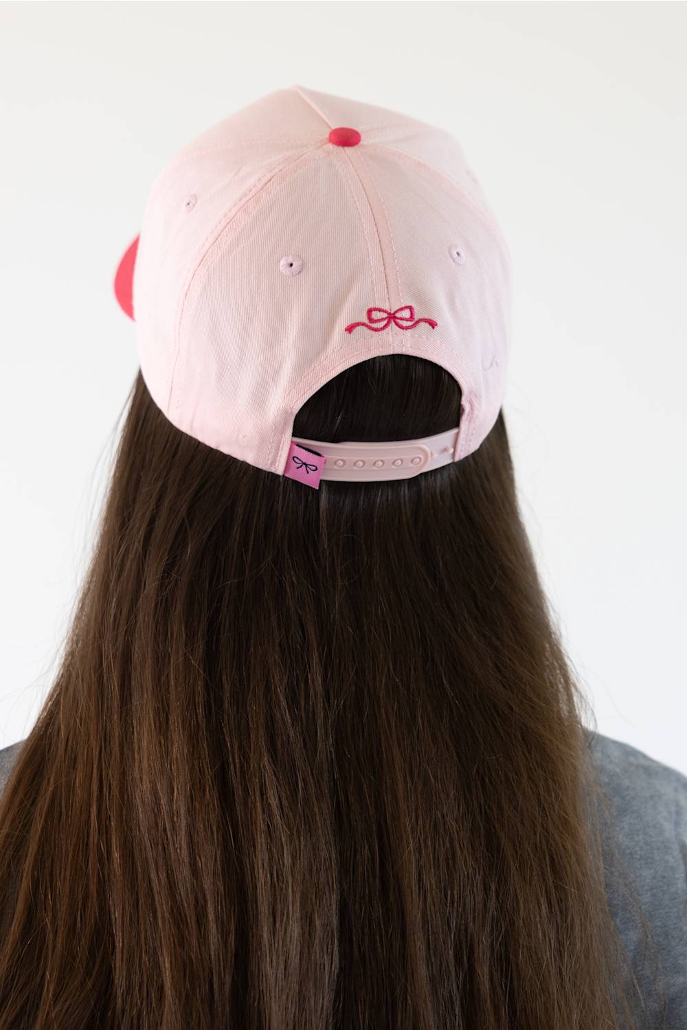 A light pink baseball cap with a small pink bow embroidery on the back, shown from behind on a model with long brown hair against a plain white background.