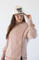 A woman wearing a dusty pink asymmetrical sweatshirt and a cream trucker hat with black text reading 'HOWDY ALL' and a brown patterned brim, posing against a white studio background.