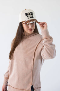 A woman wearing a dusty pink asymmetrical sweatshirt and a cream trucker hat with black text reading 'HOWDY ALL' and a brown patterned brim, posing against a white studio background.