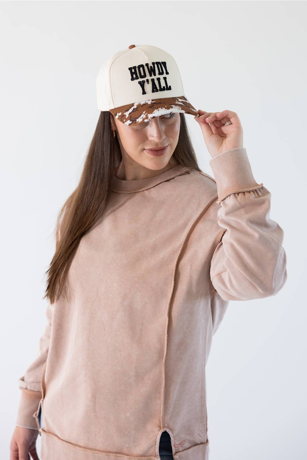 A woman wearing a dusty pink asymmetrical sweatshirt and a cream trucker hat with black text reading 'HOWDY ALL' and a brown patterned brim, posing against a white studio background.
