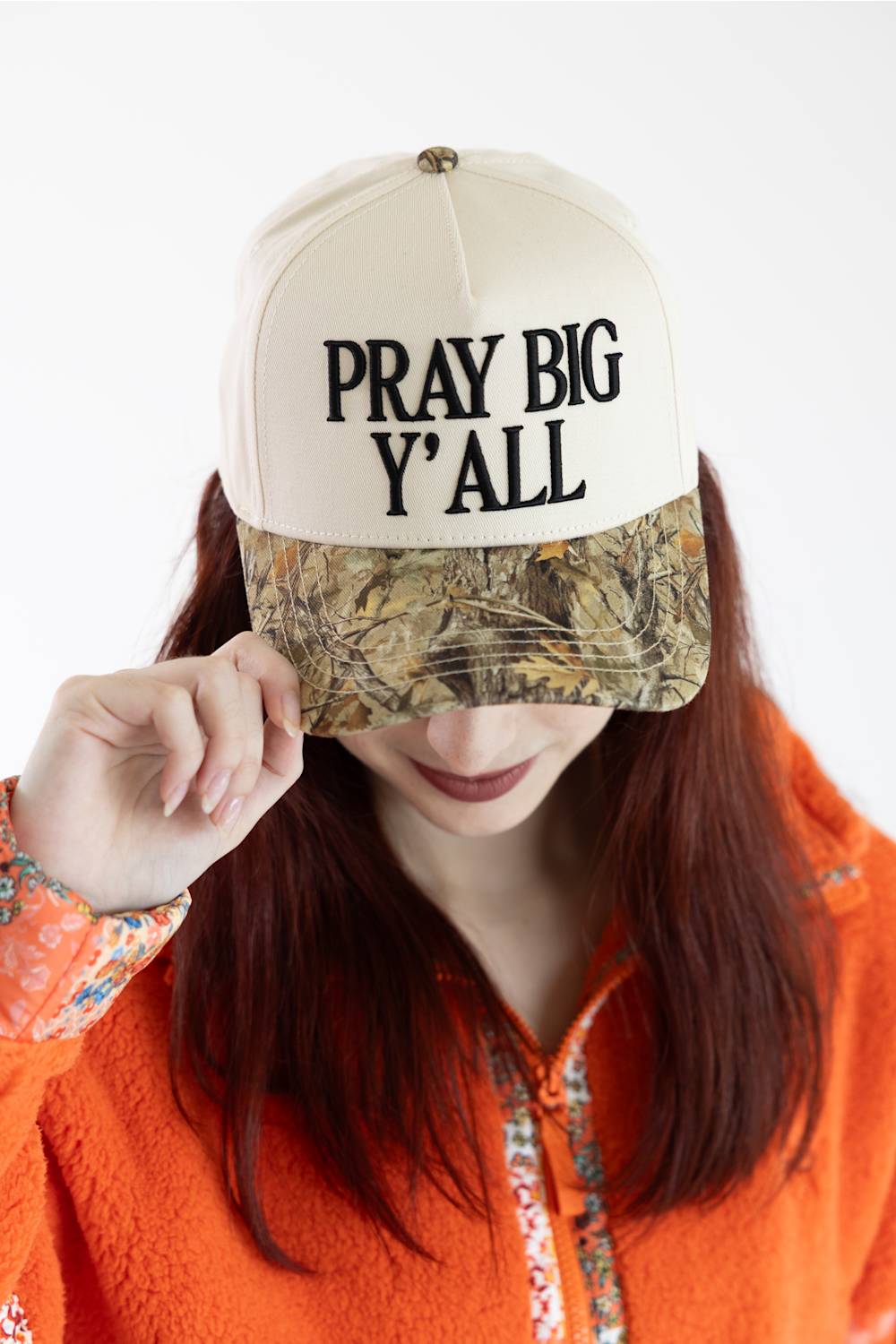 A woman wearing a beige baseball cap with black embroidered text reading 'PRAY BIG Y'ALL' and a camouflage-patterned visor, paired with an orange fleece hoodie against a plain white background.