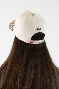 Back view of a person with long brown hair wearing a cream-colored baseball cap with a leopard-print side panel and black embroidered bow detail on the back.