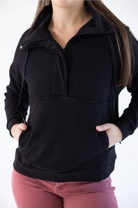 A woman wearing a black ribbed quarter-zip hoodie with a stand-up collar and front pockets, paired with dusty rose pants, against a plain white background.