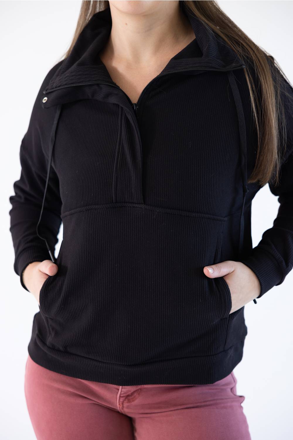 A woman wearing a black ribbed quarter-zip hoodie with a stand-up collar and front pockets, paired with dusty rose pants, against a plain white background.