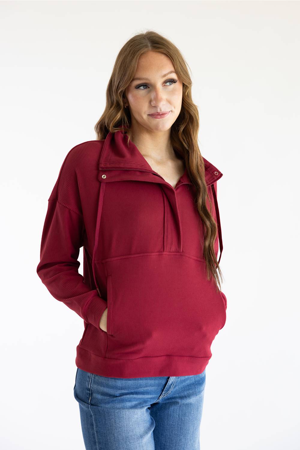 A woman modeling a burgundy half-zip hoodie with a drawstring collar and front pouch pocket, paired with blue jeans, against a plain white background.