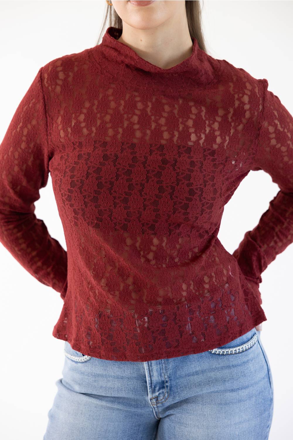 A woman wearing a long-sleeve, mock-neck lace top in a deep burgundy red, paired with light blue jeans, shown against a plain white background.