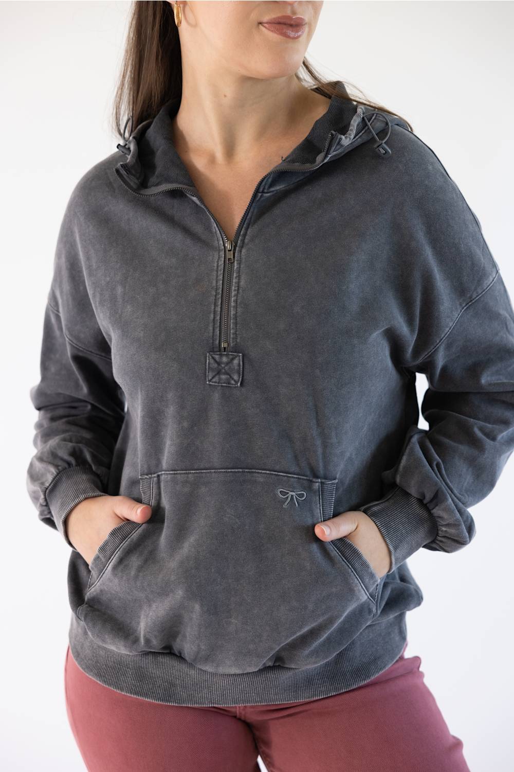 A woman wearing a dark gray washed half-zip hoodie with a drawstring hood and front kangaroo pockets, paired with dusty rose pants, against a plain white background.
