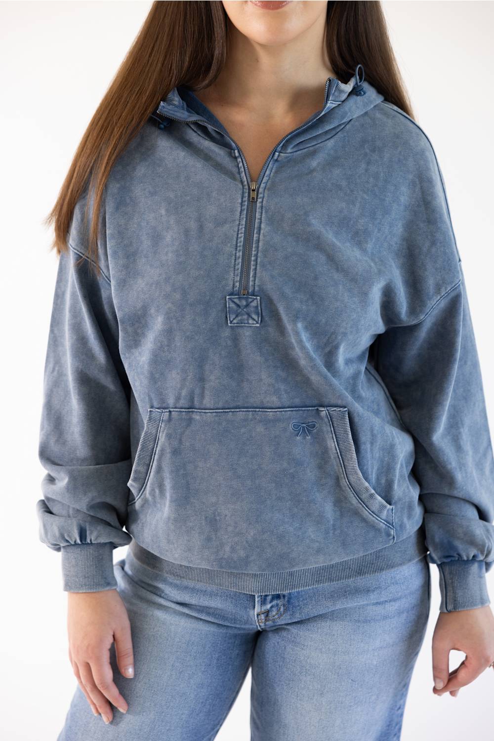 A woman wearing a washed blue half-zip hoodie with a kangaroo pocket and matching blue jeans, shown from the neck down against a plain white background.