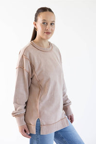 A woman modeling a light beige long-sleeve crewneck sweatshirt with unique diagonal seam details and rolled cuffs, paired with blue jeans against a plain white studio background.