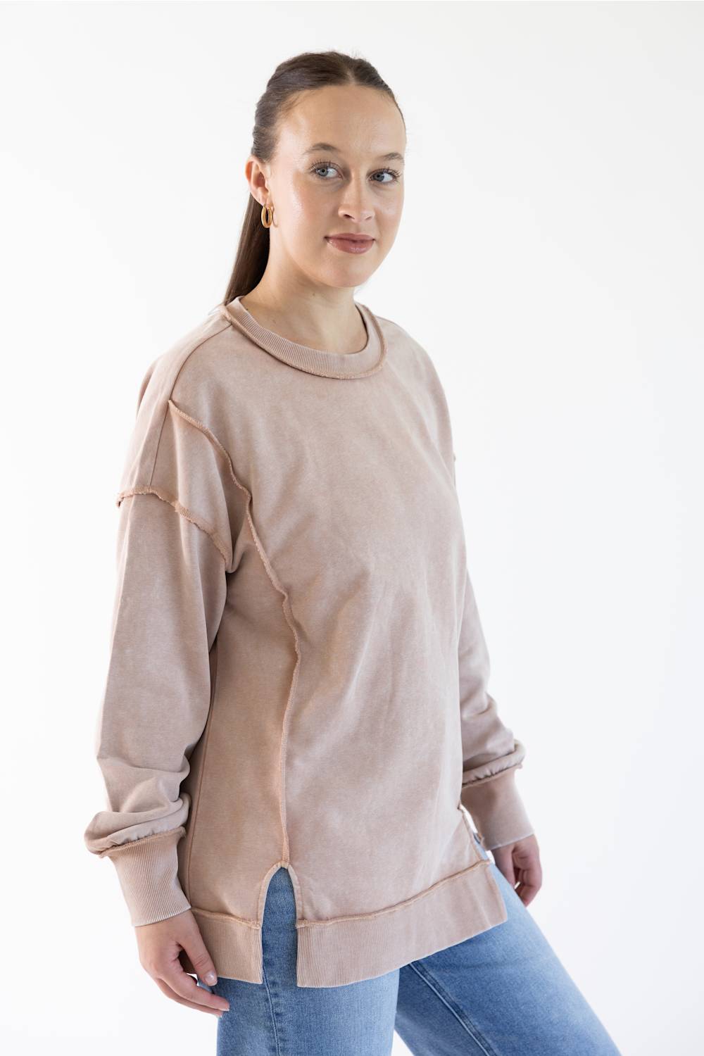 A woman modeling a light beige long-sleeve crewneck sweatshirt with unique diagonal seam details and rolled cuffs, paired with blue jeans against a plain white studio background.