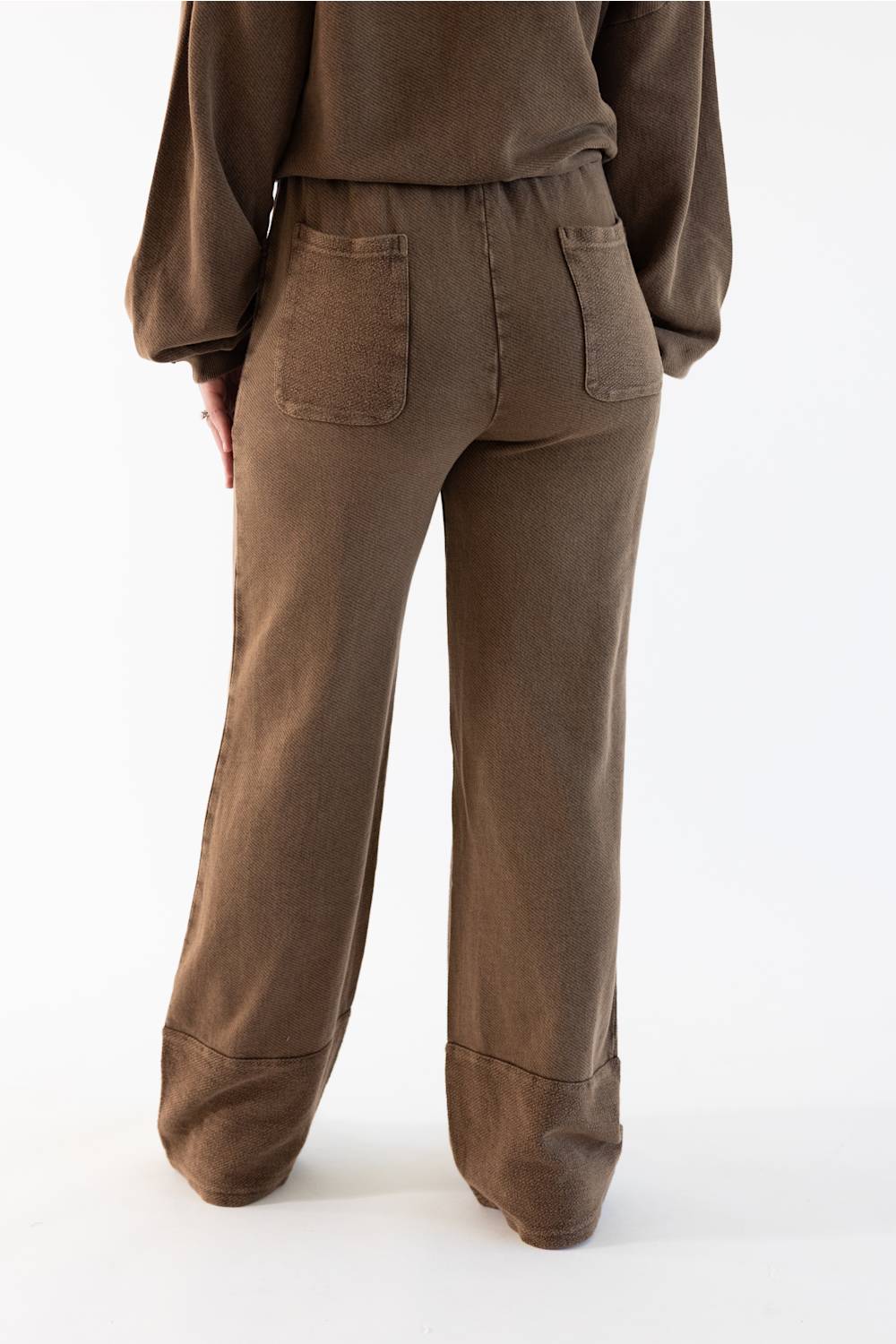 A person shown from the back wearing loose brown wide-leg pants with a matching brown top, photographed against a clean white studio background.
