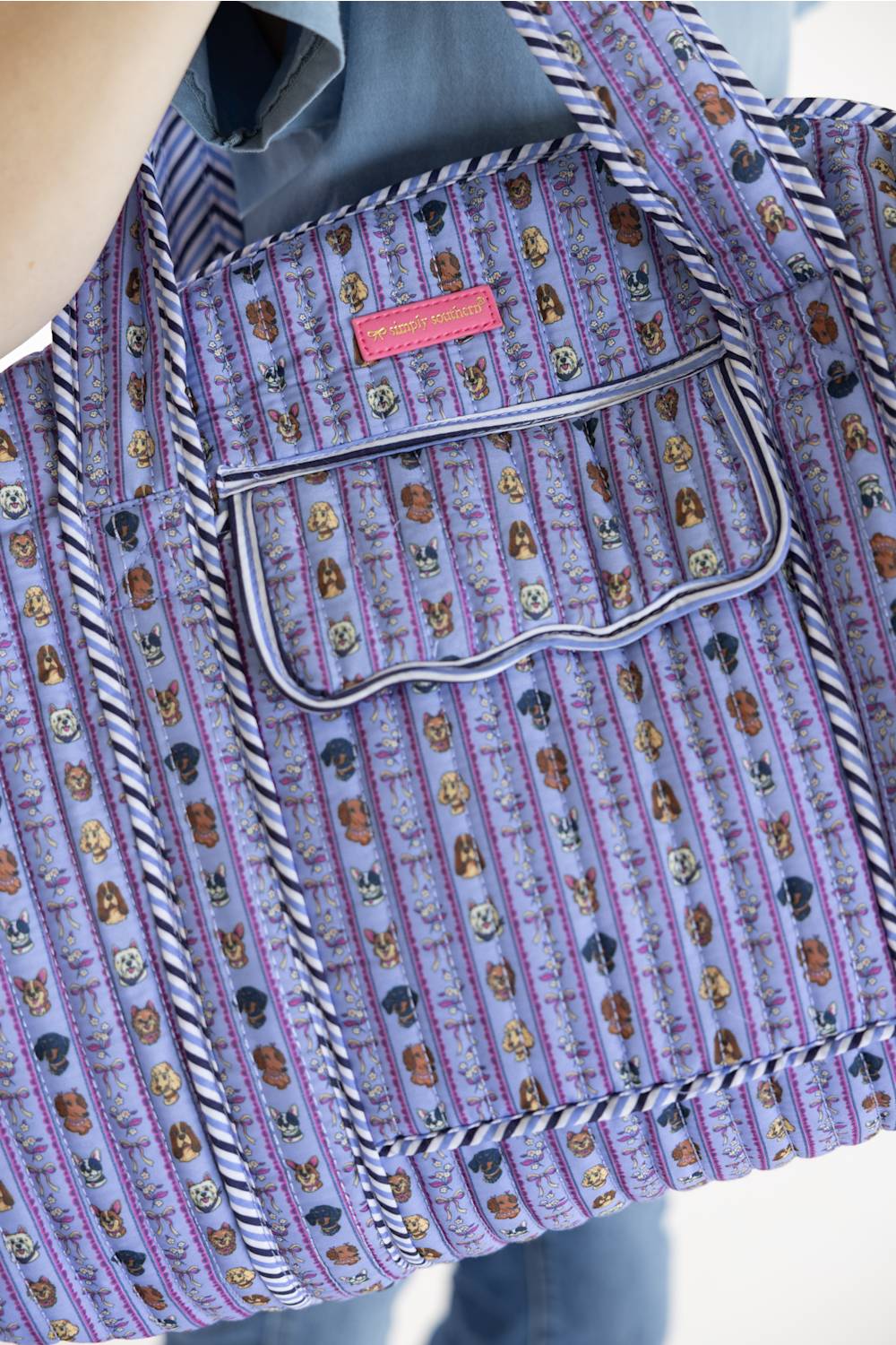 A close-up of a quilted tote bag with a purple and lavender background, covered in a repeating pattern of small dog faces. The bag features thick braided straps, a front pocket with a scalloped edge, and a small pink brand label.