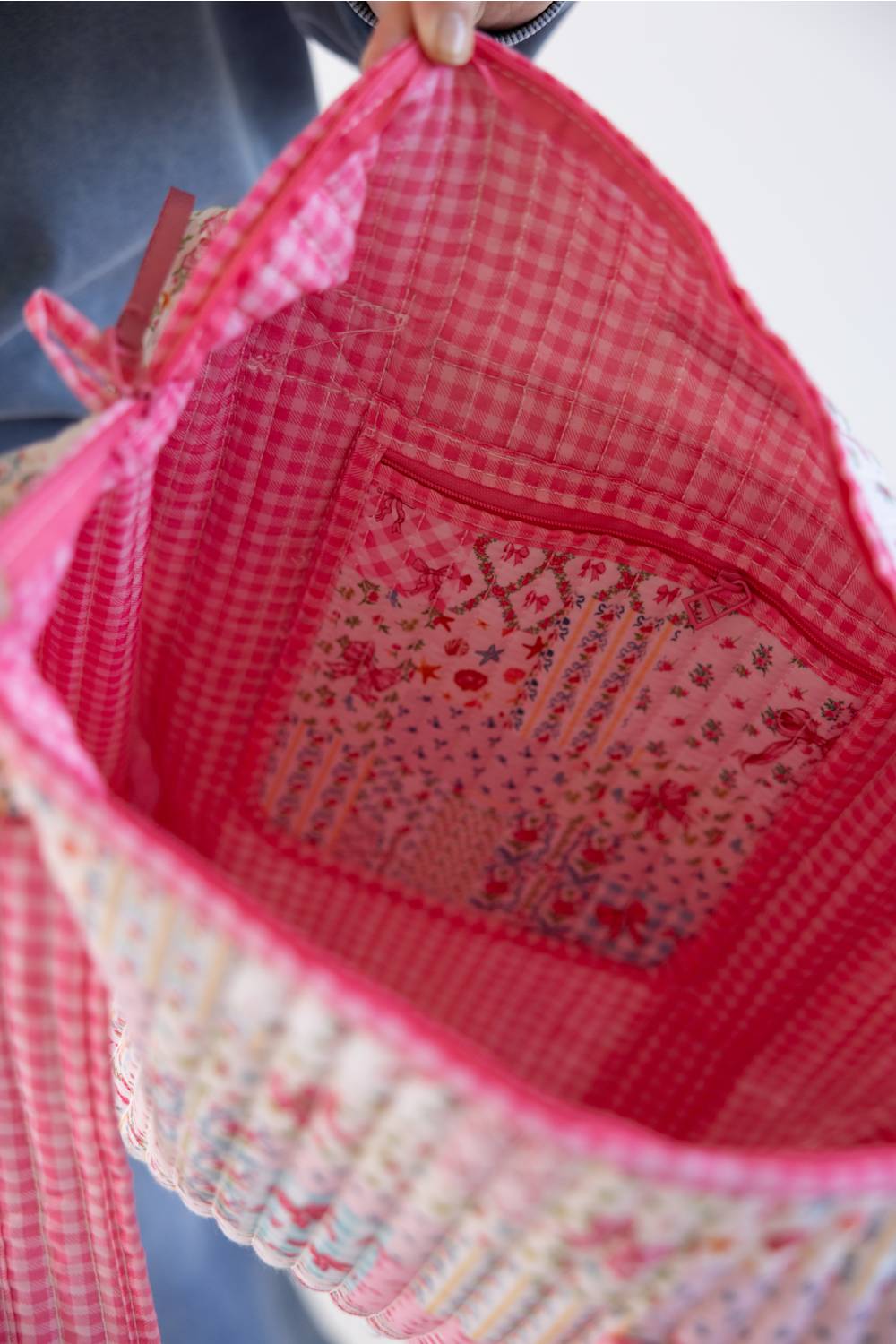 Close-up of the inside of a pink and red gingham fabric tote bag, showing a floral patterned lining and a red zipper pocket, with a hand holding the top edge.