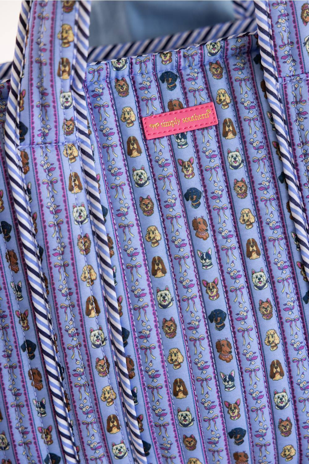 A close-up of a patterned shirt fabric with vertical stripes of navy and white, featuring cartoon dog illustrations on a light purple background. A pink brand label reading 'Simply Southern is visible near the collar.