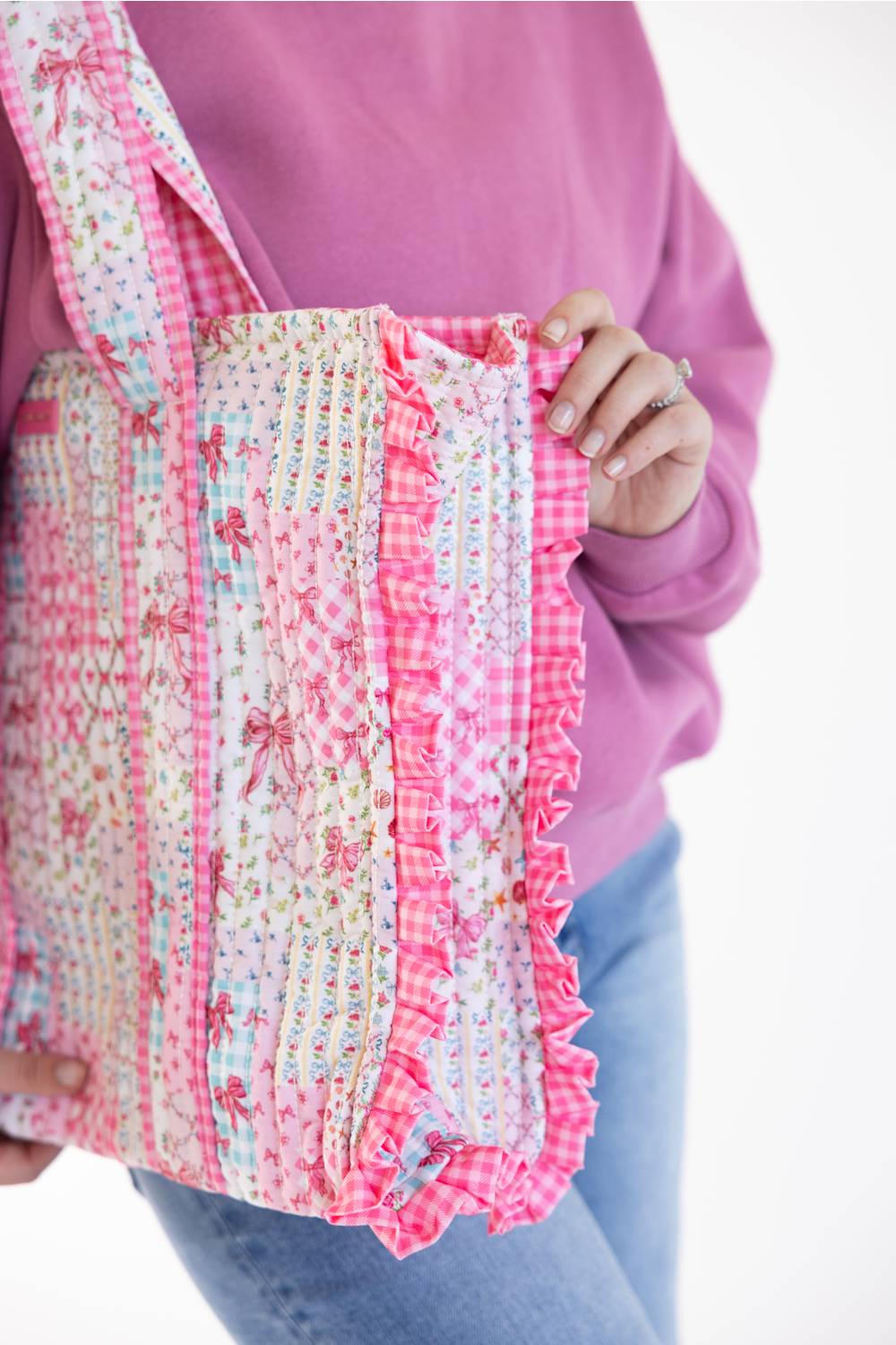 A person wearing a pink sweatshirt and blue jeans holds a large patchwork fabric tote bag with pink gingham, floral prints, and ruffled trim against a white background.