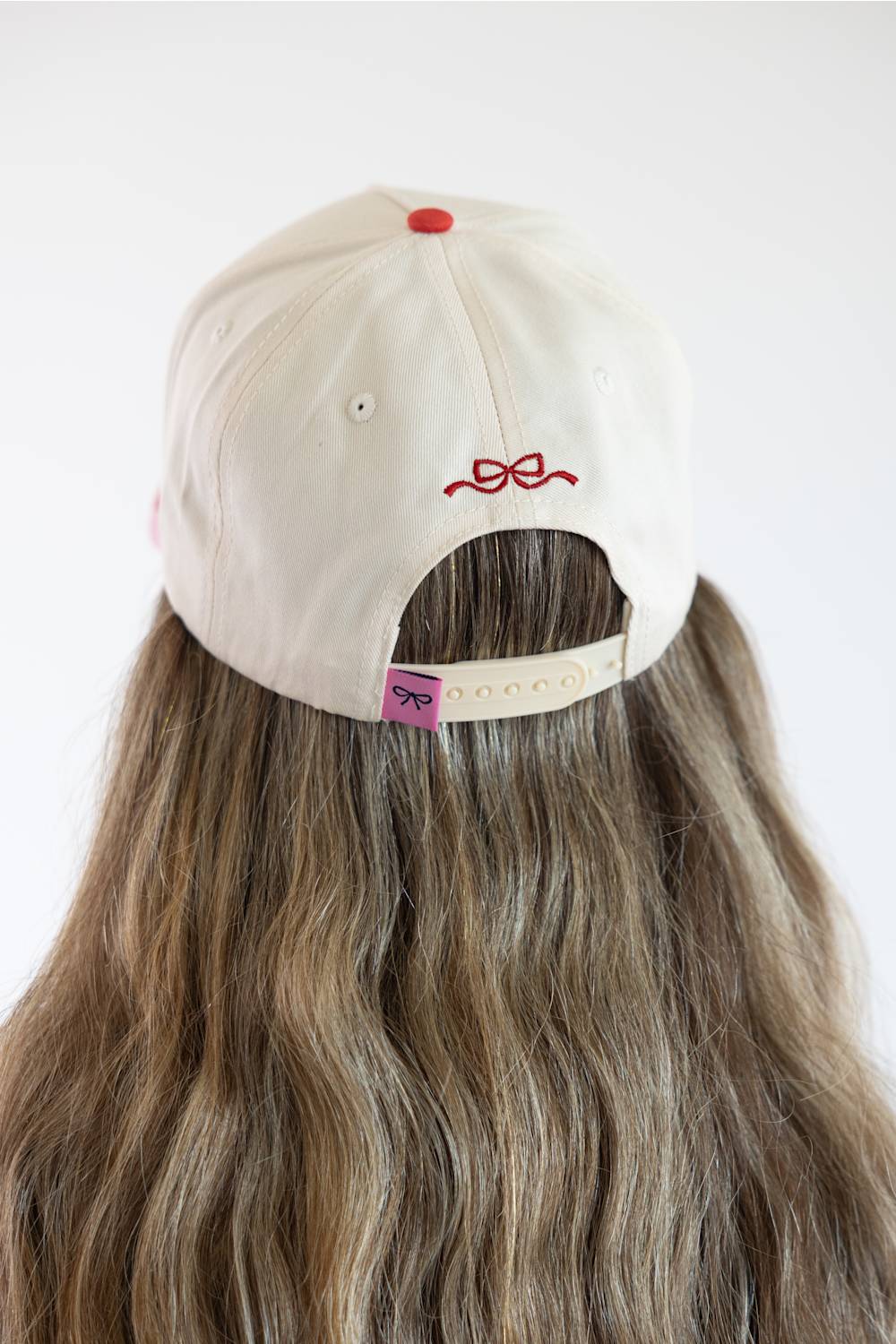 Back view of a model wearing the Simply Southern Santa Dogs Holiday Hat for Women in Cream and Red/Pink Stripe, featuring a red embroidered bow above the keyhole cutout and a pink bow tab on the adjustable cream snapback closure.