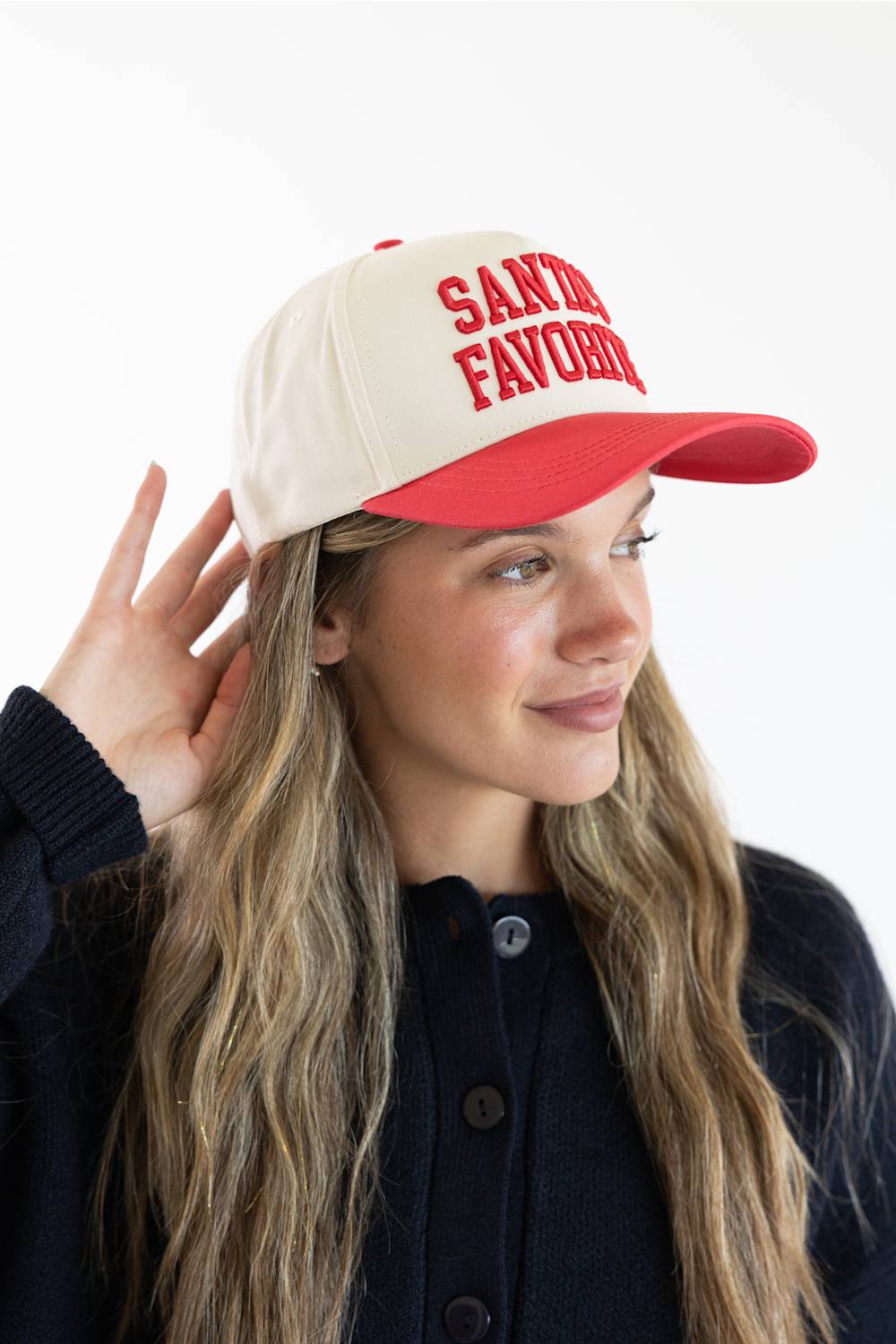 Side-profile view of a smiling model touching her hair while wearing the Simply Southern Santa’s Favorite Holiday Hat for Women in Cream/Red, featuring red embroidered collegiate-style lettering reading “SANTA’S FAVORITE” on the cream front panel and a solid red visor, paired with a navy button-down sweater against a dark background.