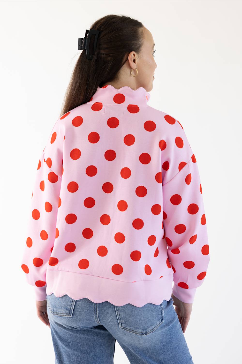 A woman wearing a pink long-sleeve quarter-zip pullover sweatshirt with large red polka dots and scalloped edge, paired with blue jeans, against a plain white background.