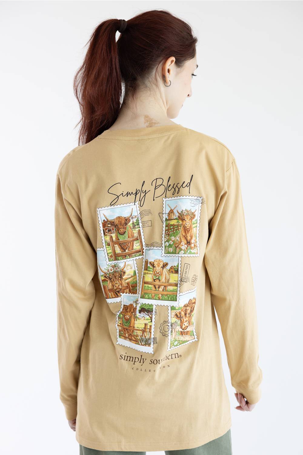 A woman shown from the back wearing a beige long-sleeve t-shirt with a large graphic print of cows and the text 'Simply Blessed' on the back, set against a plain white background.