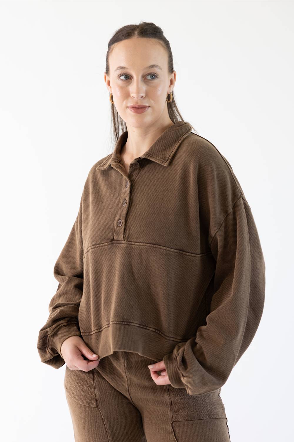 A woman modeling a brown long-sleeve polo-style sweatshirt with a button placket and matching brown pants against a plain white studio background.