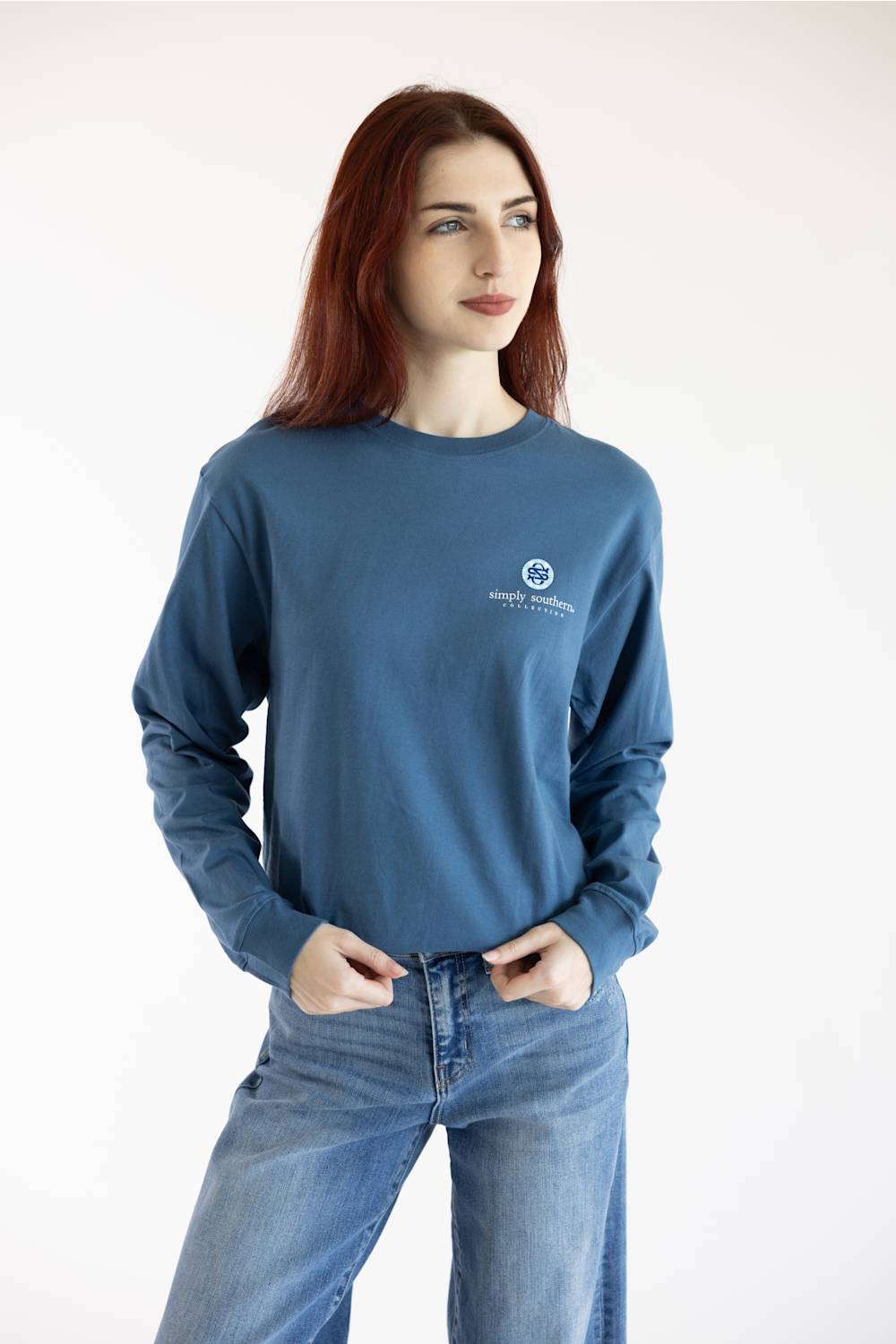 A woman with red hair wearing a slate blue long-sleeve crewneck sweatshirt with a small white circular logo on the left chest, paired with blue jeans, against a plain white studio background.