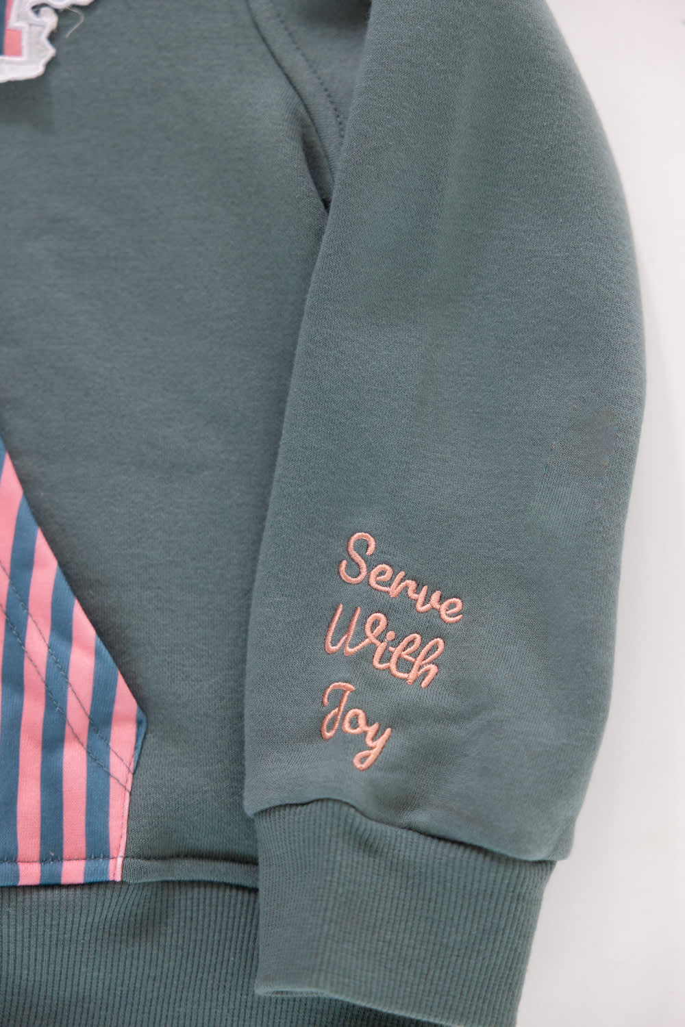 Close-up shot of the sleeve cuff on a girls' gray pullover hoodie featuring pink embroidered cursive text reading “Serve With Joy”, with a portion of the pink and navy striped kangaroo pocket visible to the left.