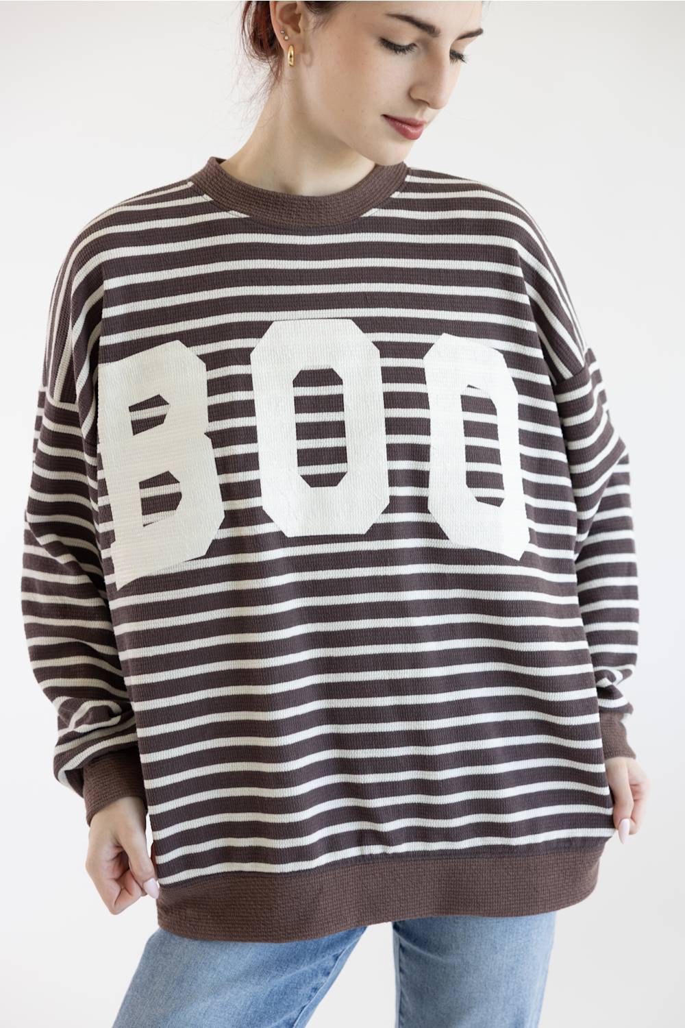 A woman posing in a brown and white horizontally striped sweatshirt with a large white BOO graphic on the front, paired with light blue jeans, against a plain white background.