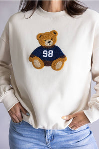 A person wearing a cream-colored crewneck sweatshirt with a teddy bear graphic on the chest. The bear is brown and wears a navy blue jersey with the number 98.