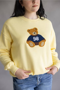 A woman wearing a pale yellow crewneck sweatshirt with a knitted teddy bear graphic on the chest wearing a navy blue jersey with the number 98, paired with blue jeans.