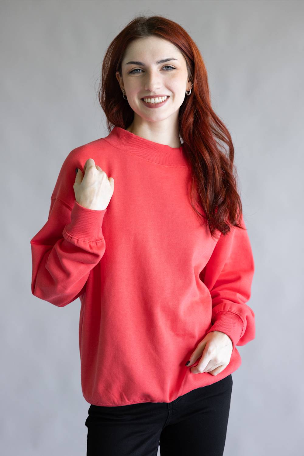 A smiling young woman with long red hair wearing a bright coral-red crewneck sweatshirt and black pants, standing against a plain gray studio background.