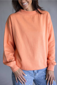 A woman wearing a peach-colored crewneck sweatshirt and light blue jeans, shown from the waist up against a neutral gray background.