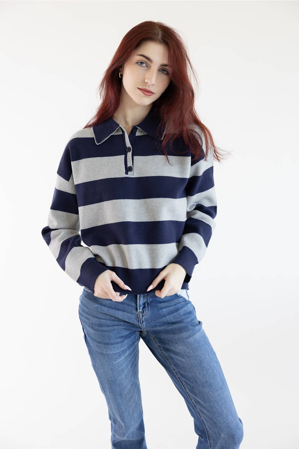 A woman with red hair wearing a navy and gray horizontally striped long-sleeve polo sweater and blue jeans, standing against a plain white studio background.