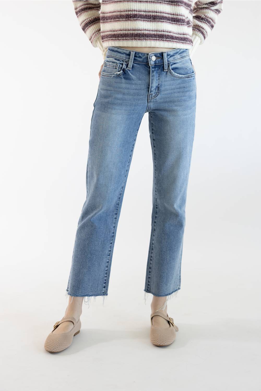 A person wearing light blue cropped jeans with a straight-to-medium leg, paired with a striped knit sweater and beige flat shoes, shown against a plain white studio background.