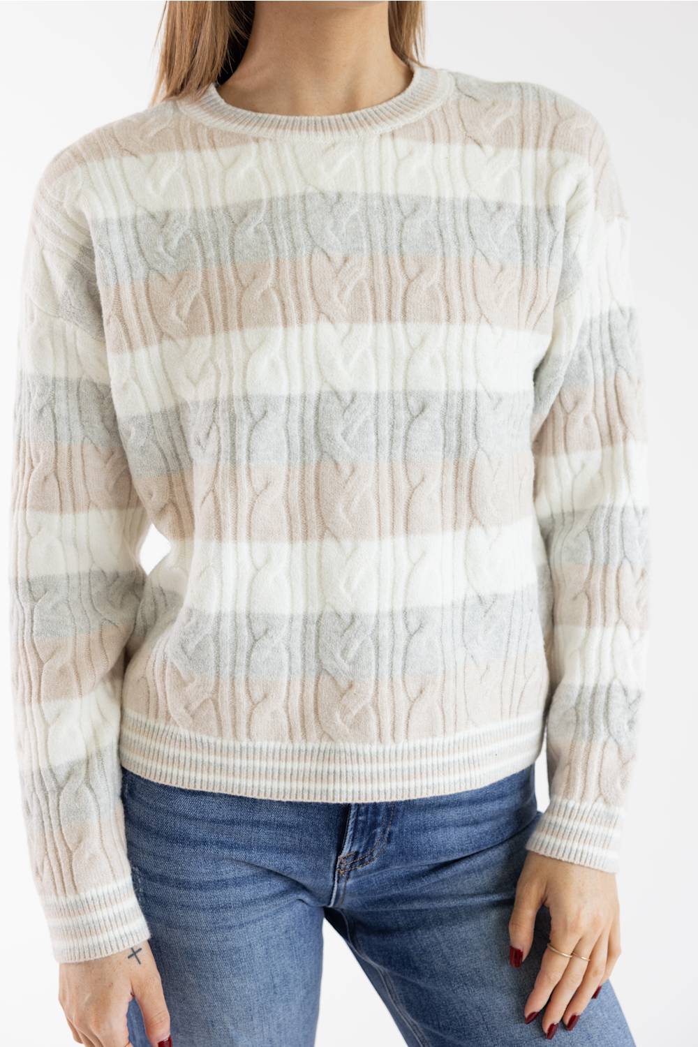 A woman wearing a cream, beige, and light gray striped cable-knit sweater with blue jeans, shown from the neck down against a plain white background.