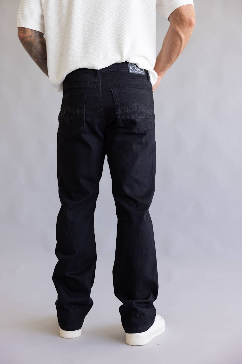 A person shown from the back wearing dark indigo denim jeans, a white textured t-shirt, and white sneakers against a light gray studio background.