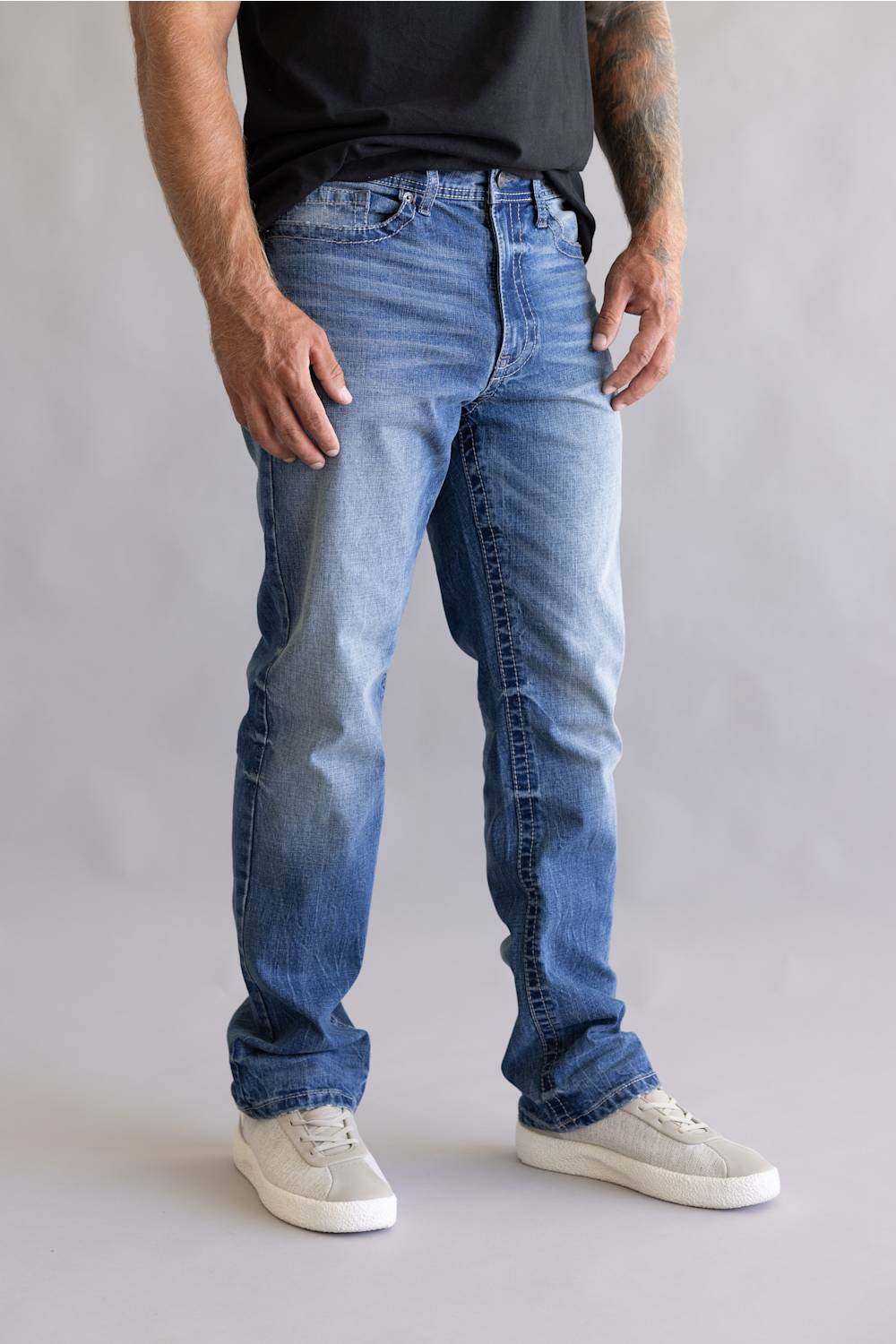 A person wearing light-to-medium wash blue jeans with a faded front thigh, paired with a black t-shirt and white sneakers, standing against a neutral gray studio background.